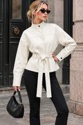 A woman wearing a cream-colored faux leather belted jacket with a mock neck, paired with black pants, sunglasses, and a black handbag, standing on a city street.