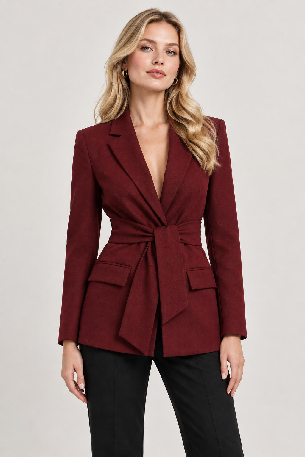 A woman modeling a structured blazer in deep burgundy with a wrap-style front and flap pockets, paired with black pants, against a plain light gray studio background.